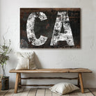 Personalized USA state location wall art featuring your custom two-letter state abbreviation on a dark, distressed industrial farmhouse background with vintage rustic charm and faded white lettering. Be surrounded by decor you LOVE! Choose between premium framed canvas or durable outdoor metal. Toe Fish Art makes artisan wall art with American materials. Proudly Made in the USA, each piece is expertly constructed with genuine craftsmanship, heritage quality, and rustic charm.